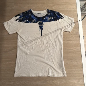 Vit t-shirt från Marcelo Burlon  - Snygg vit t-shirt med korta ärmar och ett fett blått fågeltryck över axlarna och halsen. Rund halsringning och normal passform. Materialet känns som mjuk bomull, perfekt för chill dagar. Trycket ger en unik vibe och sticker ut på ett stilrent sätt. Lite stor som du kan se, jag är 185cm och väger 75kg
