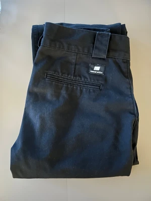 Svarta kostymbyxor från Sweet Skibs - Snygga svarta kostymbyxor från Sweet Skibs med klassisk design och diskret logga ovanför bakfickan. Byxorna har hällor för bälte och raka ben, perfekta för en clean och stilren look. Materialet känns slitstarkt och har en matt finish.