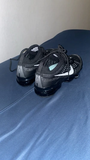 Nike Vapormax svarta sneakers - Säljer ett par svarta Nike Vapormax sneakers med vit swoosh på sidan. Skorna har stickad ovandel i syntetmaterial och genomskinlig, bubblig sula som ger en futuristisk vibe. Klassisk snörning och låg profil gör dem perfekta för dig som gillar streetwear.