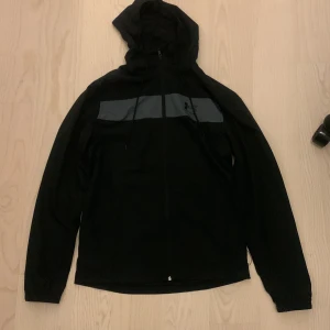 Svart vindjacka från Under Armour - Svart vindjacka från Under Armour med huva och dragsko. Jackan har en grå panel över bröstet med logga, hel dragkedja framtill och elastiska muddar vid ärmsluten. Perfekt för dig som gillar sportig och clean stil. Helt ny och aldrig använd 