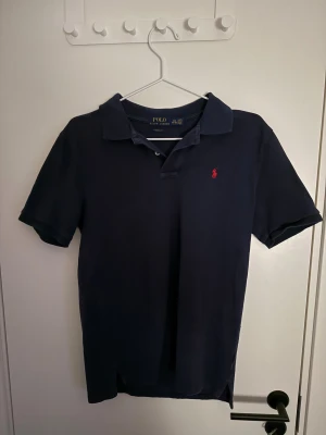 Mörkblå pikétröja från Polo Ralph Lauren - Snygg mörkblå pikétröja från Polo Ralph Lauren med klassisk passform och korta ärmar. Tröjan har krage, knappar framtill och den ikoniska röda Polo-loggan broderad på bröstet. Tillverkad i mjuk bomull som är skön mot huden.