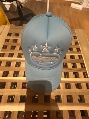Ljusblå Corteiz truckerkeps med mesh - Säljer en ljusblå truckerkeps från Corteiz med mesh på sidorna och bak. Kepsen har broderade vita stjärnor och ett broderat motiv framtill. Justerbar snapback och broderad Corteiz-logga bak. Perfekt för dig som vill ha streetwear-vibe. Den har några få fläckar på sig men inga stora skador. Priser går att diskutera