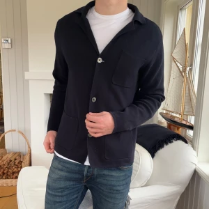 Svart cardigan jacka från Bläck - Svart cardigan jacka från Bläck i storlek M. Jackan har tre framfickor, klassisk krage och stängs med knappar. Materialet är stickat och ger en chill och stilren vibe, perfekt att slänga på över en t-shirt. Enkel och snygg design som funkar till jeans.