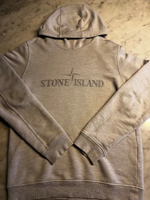 Stone Island hoodie - Snygg grå hoodie från Stone Island med broderad logga på bröstet. Tröjan har huva, långa ärmar och ribbade muddar vid ärmslut och nederkant. Perfekt för dig som gillar stilrena och klassiska streetwear-plagg.