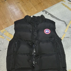 Svart dunväst från Canada Goose - Svart dunväst från Canada Goose med klassisk logga på bröstet och broderad gul etikett på insidan. Västen har hög krage, tryckknappar och dragkedja framtill samt två stora fickor. Perfekt för kyliga dagar och enkel att matcha med olika outfits.