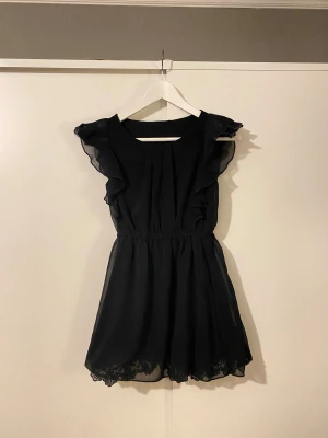 Klänning med volang och spets - Kort klänning med volang i axlar, spets under yttersta tyget i axlar och nertill. Resår i midjan