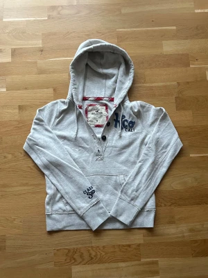 Grå cardigan från Hollister - Säljer en grå cardigan från Hollister med blått broderat Hco Cali på bröstet och tryck på ärmen. Jackan har huva med snörning, knappslå och ficka framtill. Insidan av huvan har röd och vit rutig detalj. Perfekt för chill dagar.