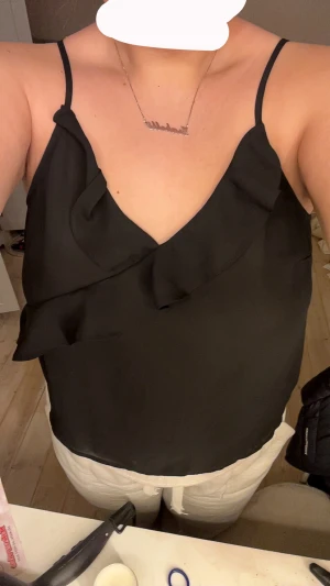Svart linnetopp med volang - Säljer en svart linnetopp med smala axelband och snygg volangdetalj framtill. Toppen har en djup v-ringning och är tillverkad i ett lätt, mjukt material som ger en skön känsla. Perfekt för dig som gillar stilrena och trendiga plagg.