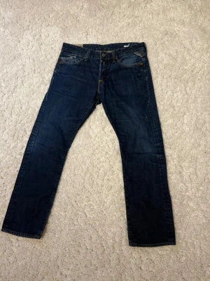 Replay - Säljer ett par replay jeans i mörkblå tvätt med klassisk straight fit. Jeansen har fem fickor, snygga kontrastsömmar och replay patch bak i midjan. Perfekt för dig som gillar tidlös stil och vill ha ett par jeans som funkar till allt.