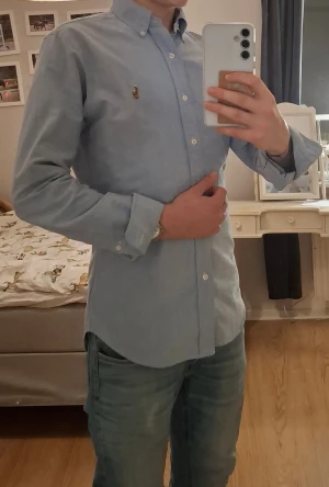 Blå slim fit skjorta Ralph Lauren - Snygg Blå skjorta från Ralph Lauren med Slim fit  passform och button-down krage. Skjortan har långa ärmar, vita knappar och den ikoniska broderade loggan på bröstet. Tillverkad i mjuk bomull som känns skön mot huden. Perfekt för en clean och stilren look.