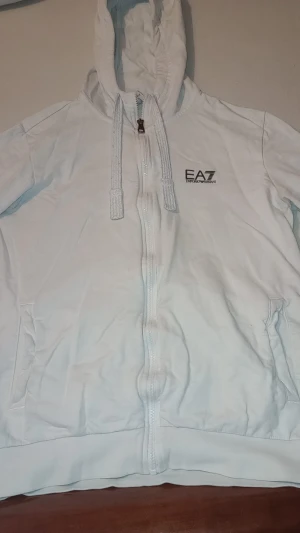 Vit hoodie EA7 Emporio Armani M - Snygg vit hoodie från EA7 Emporio Armani i storlek M. Tröjan har huva med snörning, dragkedja framtill och två fickor på sidorna. Loggan är tryckt på bröstet. Mjuk och skön bomullskvalitet, perfekt för chill eller streetwear. Och använd två gånger. 