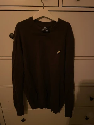 Olivgrön stickad tröja Lyle & Scott - Snygg olivgrön stickad tröja från Lyle & Scott i storlek S. Klassisk rund hals och långärmad modell med den ikoniska gula fågeln broderad på bröstet. Perfekt för dig som gillar stilrena och tidlösa plagg med en modern touch.