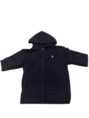 Svart hoodie från Polo Ralph Lauren - Svart hoodie med dragkedja från Polo Ralph Lauren. Klassisk design med huva och dragsko, två fickor fram och broderad logga på bröstet. Tillverkad i mjukt bomullsmaterial som är skönt mot huden. Perfekt för chill dagar och streetwear-stil.