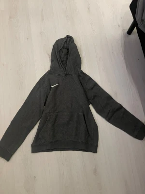 Mörkgrå Nike hoodie med huva - Snygg mörkgrå hoodie från Nike med klassisk logga på bröstet. Tröjan har huva, lång ärm och en stor magficka framtill. Mjuk insida och avslappnad passform, perfekt för chill eller träning. Materialet är bomull och polyester.