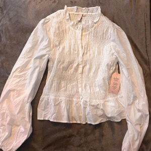 Fitted blus Nelly - Supersöt vit skjorta från Nelly i storlek 38 med smockad överdel och volangkant nedtill. Skjortan har långa ärmar, knappar framtill och en lätt rynkad struktur som ger en cool och feminin vibe. Perfekt för dig som gillar detaljer och vill sticka ut lite extra.