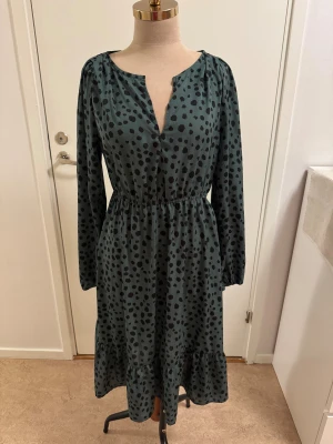 Grönprickig midiklänning från SHEIN - Grön klänning från SHEIN med svarta prickar, v-ringning och långa ärmar. Resår i midjan och volang nertill ger en avslappnad men snygg siluett. Tillverkad i mjukt syntetmaterial, perfekt för dig som gillar trendiga mönster och bekväm passform.
