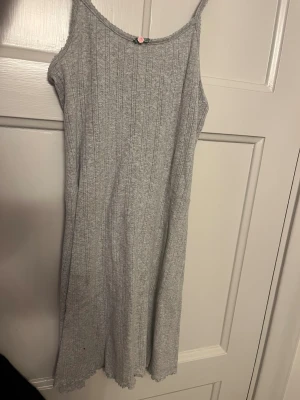 Grå ribbad kortklänning med spetskant - Söt grå kortklänning med smala axelband och ribbad struktur. Klänningen har en liten rosa blomma framtill 