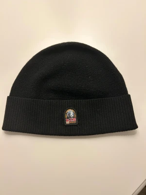 Svart mössa från Parajumpers - Snygg svart ribbstickad mössa från Parajumpers med uppvikt kant och broderad patch framtill. Mössan har även en röd etikett med logga på sidan. Perfekt för kalla dagar och enkel att matcha med din jacka.