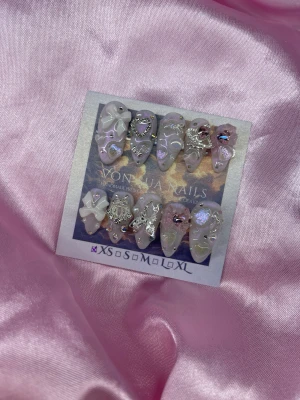 BabyShoujo (XS);Meidum Almond - Säljer ett set handgjorda press-on naglar i vitt/rosa med holografiska detaljer, 3D rosetter, hjärtan,m och silverfärgade charms. Naglarna har en medium almond-form och glittrande cateyefinish, perfekta för dig som vill ha extra glam på fingertopparna.XS: 14 mm, 11 mm, 11,5 mm, 10 mm, 8 mm 