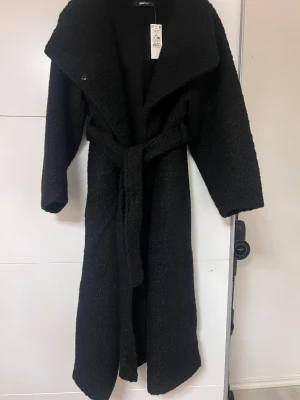 soft boucle long coat - Lång kappa i bouclé med knytskärp i midjan. Kappan är svart och har fickor i sidorna. Lång ärm och stor krage med slag. Helt ny