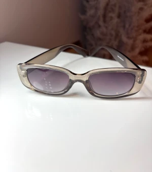 Transparenta rektangulära solglasögon - Snygga solglasögon med transparent plastbåge och rektangulär form. Glasen är tonade i grått och ger en cool vibe. Perfekt accessoar för att lyfta din stil och skydda ögonen mot solen. Det är små repor syns i bild 2. Medföljer ett fodral