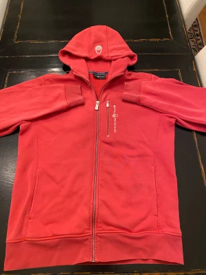 Ljus röd/ mörk rosa hoodie från Sail Racing L - Riktigt snygg ljus röd/ mörk rosa hoodie från Sail Racing med dragkedja, broderad logga på bröstet och stor Sail Racing-tryck på ryggen. Mjuk insida, två fickor fram och klassisk huva med logga. 