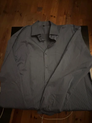 Blårandig skjorta Dressmann XL - Snygg blårandig skjorta från Dressmann i storlek XL. Skjortan har klassisk krage, knappar framtill och en bröstficka. Materialet är mjukt och känns som bomull, perfekt för dig som gillar stilrena och enkla plagg med diskret mönster.