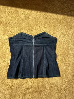 Mörkblå denim korsett-topp - Säljer en snygg mörkblå korsett-topp i denim med markerad midja och dragkedja framtill. Toppen är axelbandslös och har en cool, strukturerad form. Ända defekten är sömm som gått upp lite, men stör inte användningen. 