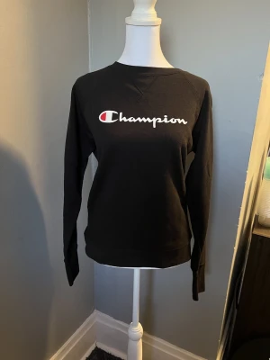 Svart sweatshirt från Champion - Svart sweatshirt från Champion med klassisk logga i vitt och rött framtill. Tröjan har rund halsringning och långa ärmar. Perfekt för en avslappnad och sportig stil.