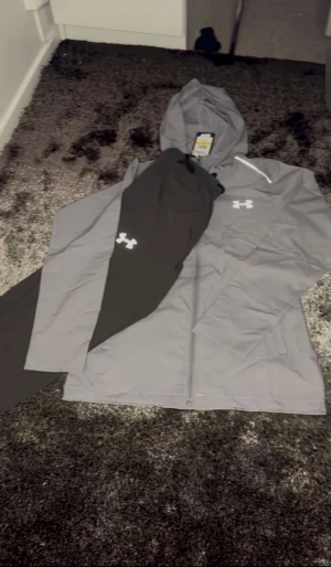 Grå vindjacka från Under Armour - Säljer en grå vindjacka från Under Armour med huva och dragkedja framtill. Jackan har en vit logga på bröstet och är tillverkad i lätt, vattenavvisande material. Perfekt för dig som vill ha en sportig och stilren look.