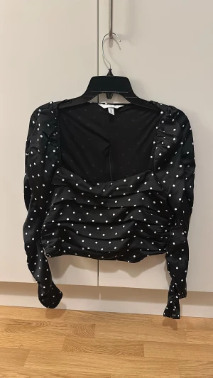 Svart prickig croppad topp H&M - Säljer en svart croppad topp från H&M med vita prickar. Toppen har långa puffärmar, rynkade detaljer framtill och baktill samt en fyrkantig urringning. Materialet är glansigt och känns lyxigt. Perfekt för dig som vill sticka ut lite extra.
