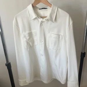 Vit Overshirt - En helt vit svart overshirt från Zara. 10/10 skick, jag köpte den förra året för 500kr, men jag har använt den 1-2 gånger. Den har inga skador eller liknande. Den är helt ny kan man säga. Väldigt bekväm, man kan ha t shirt under men även hoodie. Det är storlek L, och den sitter även som ett L plagg. Dessa säljs inte mer. Rekommenderar starkt!