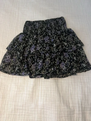 Svart blommig volangkjol med resår - Säljer en svart kortkjol med lila och vita blommor, volanger och bred resår i midjan. Kjolen har ett luftigt lager på lager-utseende och är gjord i tunn polyester med mjukt foder. Perfekt för dig som gillar romantiska detaljer och blommigt mönster. Den är stor i storlek och går att stretcha ut