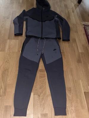 Nike Tech Fleece dress  mörkgrå - Nike Tech Fleece joggers i mörkgrå och grå färg med svart Nike-logga på benet. Byxorna har snörning i midjan och ribbade muddar vid benslut. Materialet är mjukt och skönt, perfekt för chill eller träning. Snygg tvåfärgad design med kontrastpaneler. Jätte mysig dress storlek M i mycket bra skick 