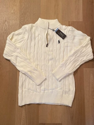 Vit stickad half zip tröja Polo Ralph Lauren - Vit stickad tröja från Polo Ralph Lauren i storlek S. Tröjan har ett klassiskt kabelstickat mönster, ribbade muddar och krage samt en half zip-dragkedja framtill. Broderad svart logga på bröstet. Perfekt för dig som gillar stilren och tidlös design.