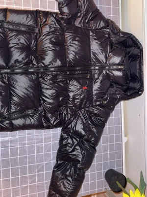 Svart pufferjacka från Polo Ralph Lauren - Säljer en svart pufferjacka från Polo Ralph Lauren med glansig finish och röd logga på bröstet. Jackan har huva, flera dragkedjefickor och är perfekt för kalla dagar. Stängs med dragkedja framtill och har långärmade ärmar. I storleken M