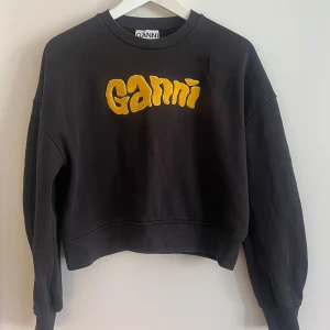 Brun Ganni sweatshirt med gul logga - Svart/mörkbrun sweatshirt från Ganni med stor gul logga framtill. Tröjan är croppad med loose passform och har långa ärmar samt ribbade muddar.                             Nypris: 1950kr