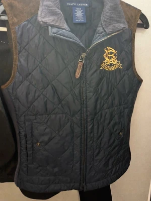 Quiltad blå väst från Ralph Lauren - Säljer en quiltad blå väst från Ralph Lauren med brunt mockadetalj på axlarna och ett broderat gult emblem på bröstet. Västen har ståkrage i manchester, dragkedja framtill och praktiska fickor med knapp. Materialet är polyester och äkta mocka.                                                                    Mycket fint skick, inga hål eller fläckar,sparsamt använd, storlek S (dam).