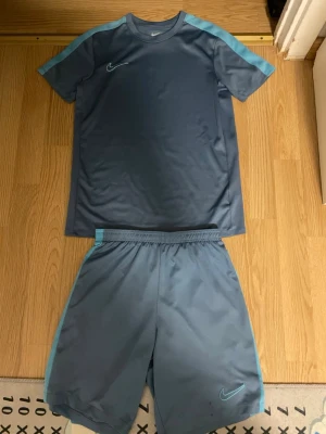 Tränings kläder och sommarkläder  - Bilderna visar ett blått Nike träningsset med t-shirt och shorts, men inga skor finns med. Setet har ljusblå detaljer och är gjort i syntetmaterial, perfekt för träning. Man kan också diskutera priset