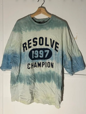 Blå tie-dye t-shirt Resolve 1997 - Säljer en oversized t-shirt från Champion med coolt tie-dye mönster i blått och vitt. Framsidan har trycket 'RESOLVE 1997 CHAMPION' i stora bokstäver. Perfekt för dig som gillar retrovibbar och streetstyle.