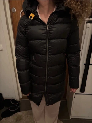 Svart lång dunjacka från Parajumpers - Säljer en svart, lång dunjacka från Parajumpers med huva och snygga detaljer som gul knapp vid halsen och patch på ärmen. Jackan har quiltad design, dragkedja framtill och är perfekt för kalla dagar. Materialet är polyester och dun, och passformen är normal., helt ny , nypris 7600 sitter som xs/s