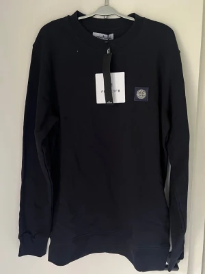 Svart sweatshirt från Stone Island - Svart långärmad sweatshirt från Stone Island med klassisk rund hals och ikonisk logopatch på bröstet. Tröjan är i mjuk bomull och har ribbade muddar vid ärmslut och nederkant. Perfekt för dig som gillar stilrena och enkla plagg med premiumkänsla.