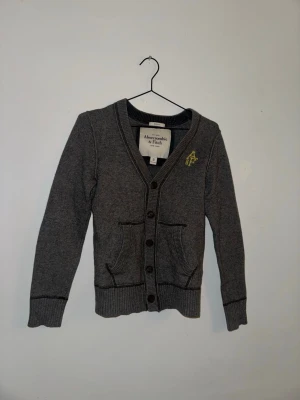 Grå kofta från Abercrombie & Fitch - Säljer en grå kofta från Abercrombie & Fitch i storlek S. Koftan har långa ärmar, ribbade muddar och knappstängning framtill. Två fickor och en gul broderad logga på bröstet ger extra stilpoäng. Perfekt för lager-på-lager under kyliga dagar.