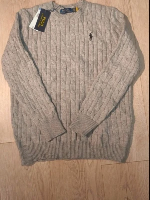 Grå stickad tröja från Polo Ralph Lauren - Säljer en snygg grå stickad tröja från Polo Ralph Lauren med klassiskt kabelmönster och broderad logga på bröstet. Tröjan har rund hals och långa ärmar med ribbade muddar. Perfekt för dig som gillar stilrena och tidlösa plagg.