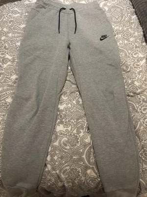 Grå Nike tech mjukis byxor - Säljer ett par grå mjukisbyxor från Nike med svart logga på vänster lår. Byxorna har svarta snören i midjan och en dragkedja på bakfickan. Sköna och stilrena, perfekta för chill eller träning. Materialet är mjukt och känns som bomull.