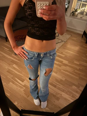 Lågmudjade blå jeans med slitningar  - Säljer ett par ljusblå bootcut jeans med stora slitningar på båda benen. Jeansen har låg midja, klassisk femficksdesign och är gjorda i jeansmaterial. Perfekta för dig som gillar en edgy och avslappnad stil.