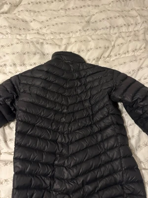 Svart Peak Performance dunjacka - Peak Performance dunjacka i svart med quiltad design och hög krage. Jackan har dragkedja framtill och två sidofickor. Materialet är syntet och dun, perfekt för kyliga dagar. Snygg logga på bröstet och diskret R&D-detalj på ärmen.