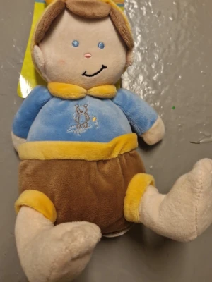 Docka med Blå plyschväst av broderi - Säljer en mjuk och gosig gossedjur i plyschmaterial med gul krage och kant. Västen har ett broderat motiv av en liten björn på framsidan och passar perfekt för dig som gillar gulliga detaljer. Materialet är syntet och färgerna är blå och gul.