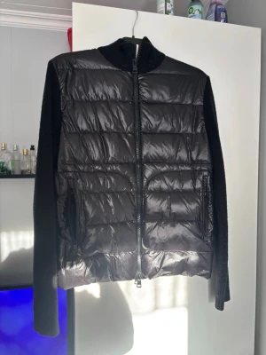 Svart pufferjacka från Moncler - Svart pufferjacka från Moncler med stickade ärmar och rygg. Framsidan är quiltad med glansigt material och har två fickor med dragkedja. Insidan av dragkedjan har en röd och vit rand. Moncler-logga på vänster ärm. Perfekt för dig som vill ha en snygg och varm jacka. Storleken är XL men sitter mer som en L. Obs jag köpte denna jacka begagnat och vet derför inte till 100% om den är äkta eller om det är en A-replika, där av kan priset diskuteras