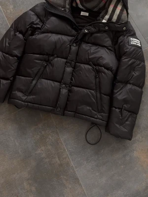 Svart pufferjacka från Burberry - Svart pufferjacka från Burberry med stor patch på ärmen och klassiskt rutigt foder i huvan. Jackan har dragkedja, knappar och två snedställda fickor med dragkedja. Perfekt för kalla dagar och har en riktigt schysst streetstil.